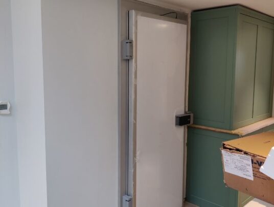 Installed walk-in cold room with a white door and green cupboard next to it.